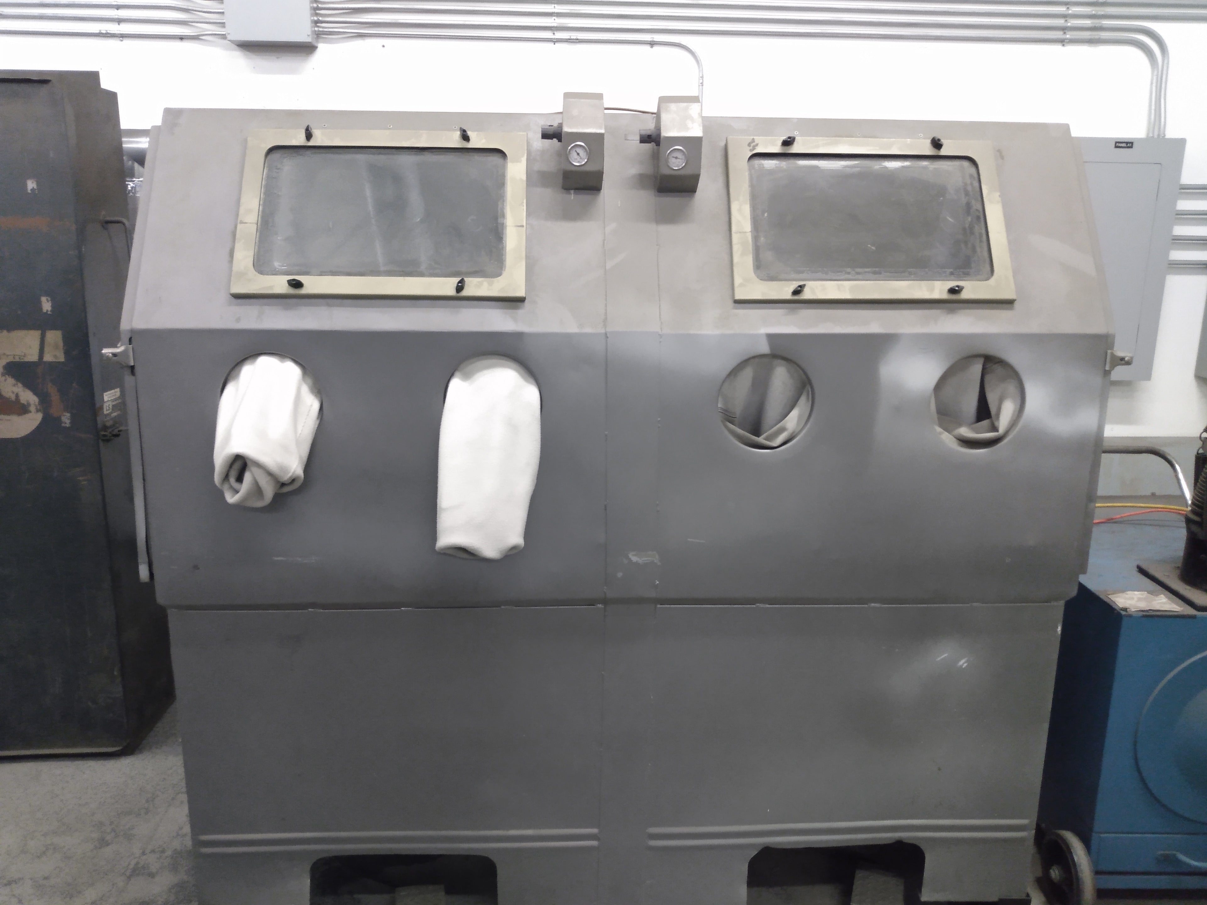 A gray metal abrasive blasting cabinet with a viewing window, internal lighting, and attached dust collection system.