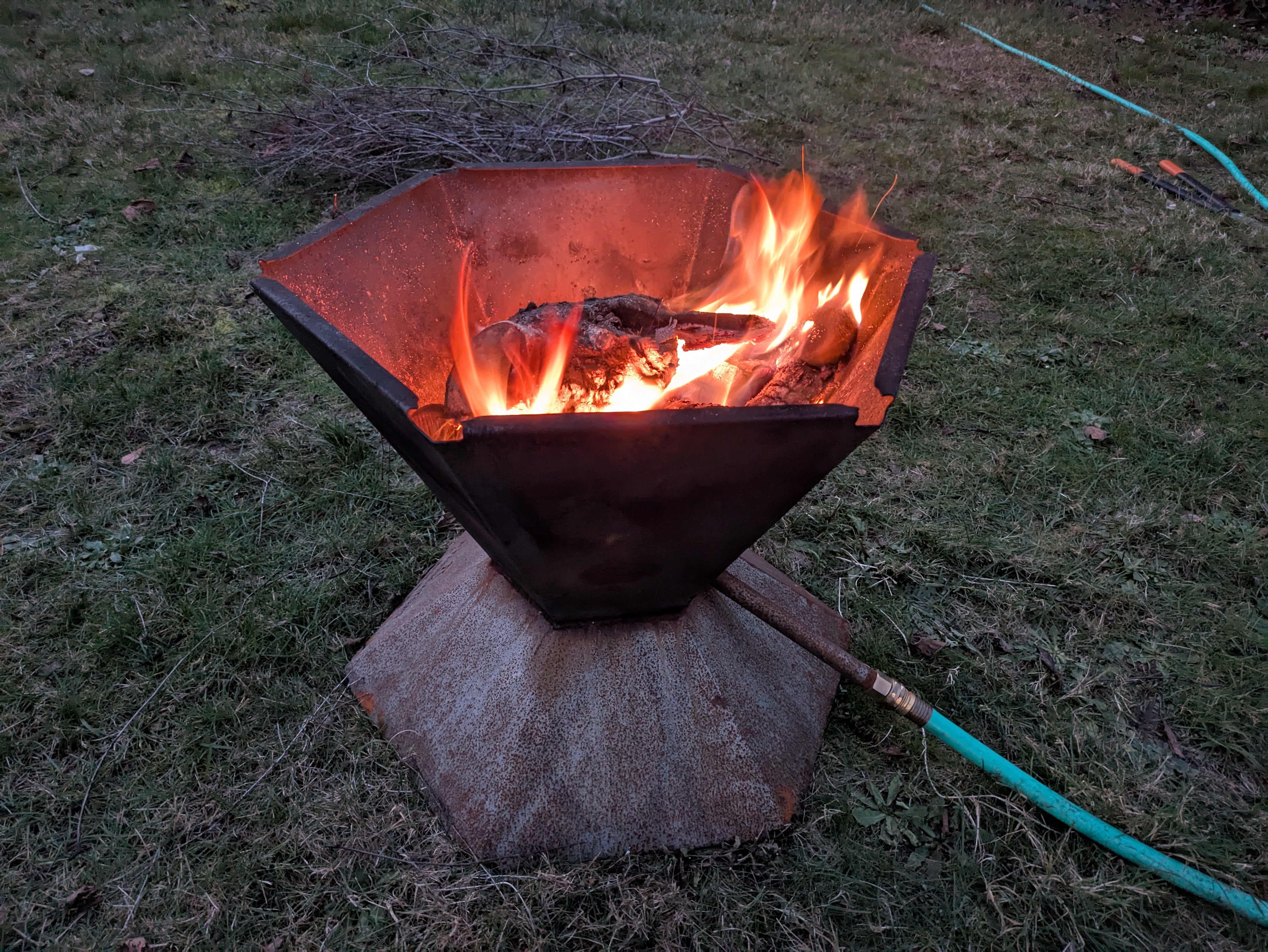 Biochar-Making Fire Pit burning wood with active flames, showing the developing patina on the cone-shaped steel body resting on its hexagonal base in a grassy yard.