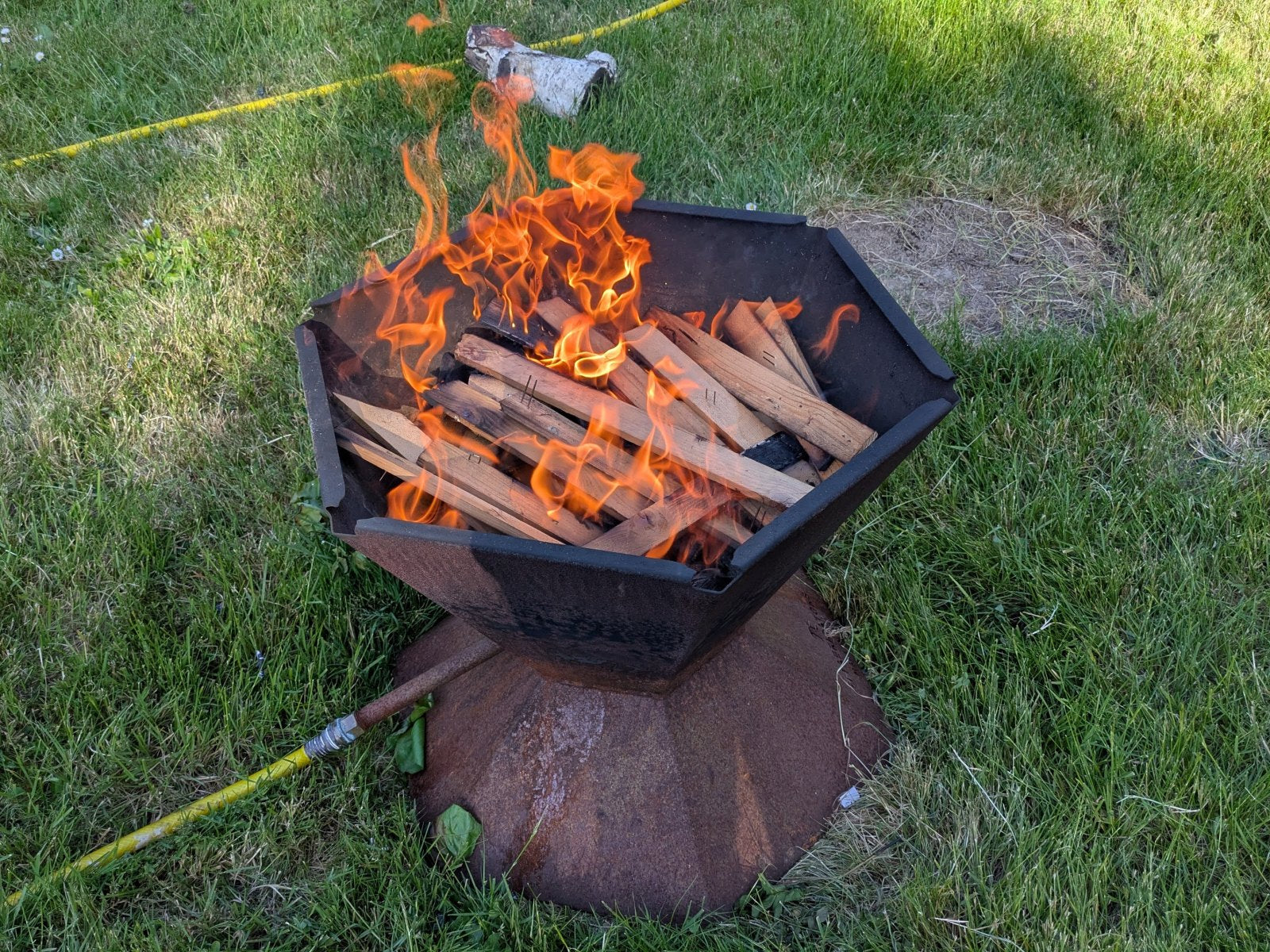A fresh layer of split wood laid flat in the Biochar-Making Fire Pit with flames coming up over it, garden hose connected to the quench pipe