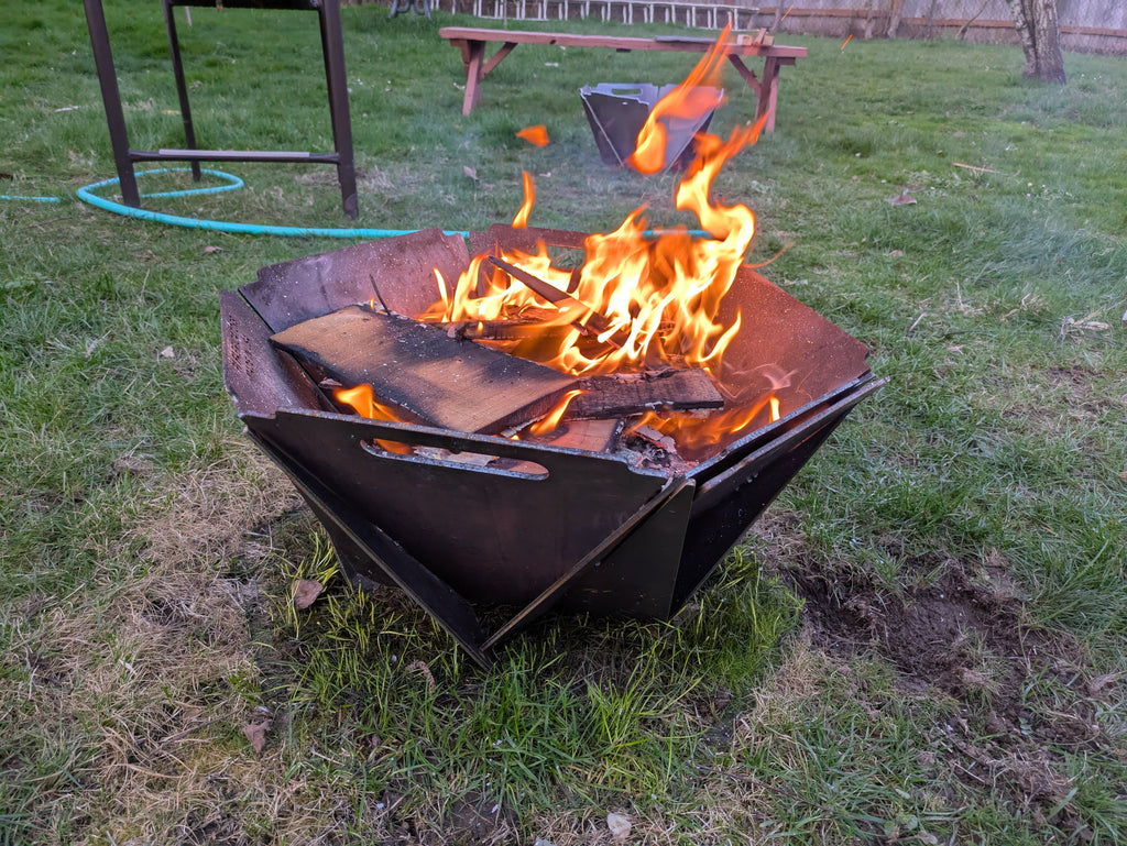 A collapsible fire pit made of steel with wood burning inside it, placed on a grassy outdoor area.