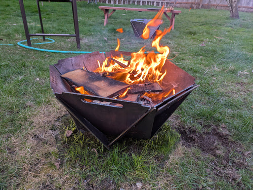 A collapsible fire pit made of steel with wood burning inside it, placed on a grassy outdoor area.
