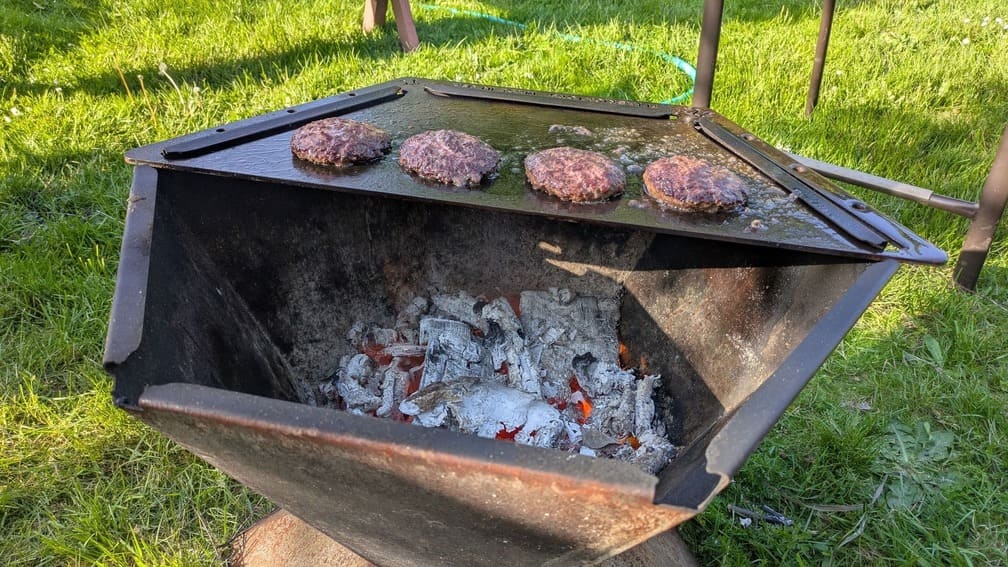 Pi Fabricators biochar kiln and hot plate grill (Made in USA) in use outdoors. Hamburgers are cooking on the grill over hot coals, with green grass surrounding the unit.