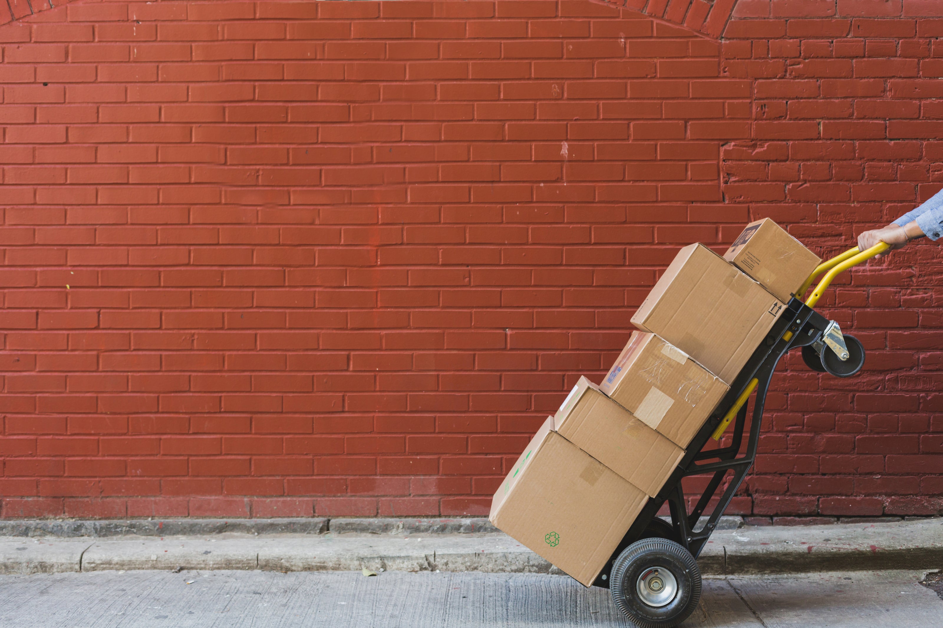 Carboard boxes stacked high on a hand truck. A red brick wall in the background. 
